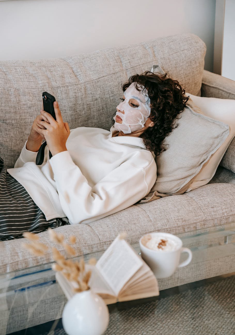 young woman in face mask using smartphone on sofa