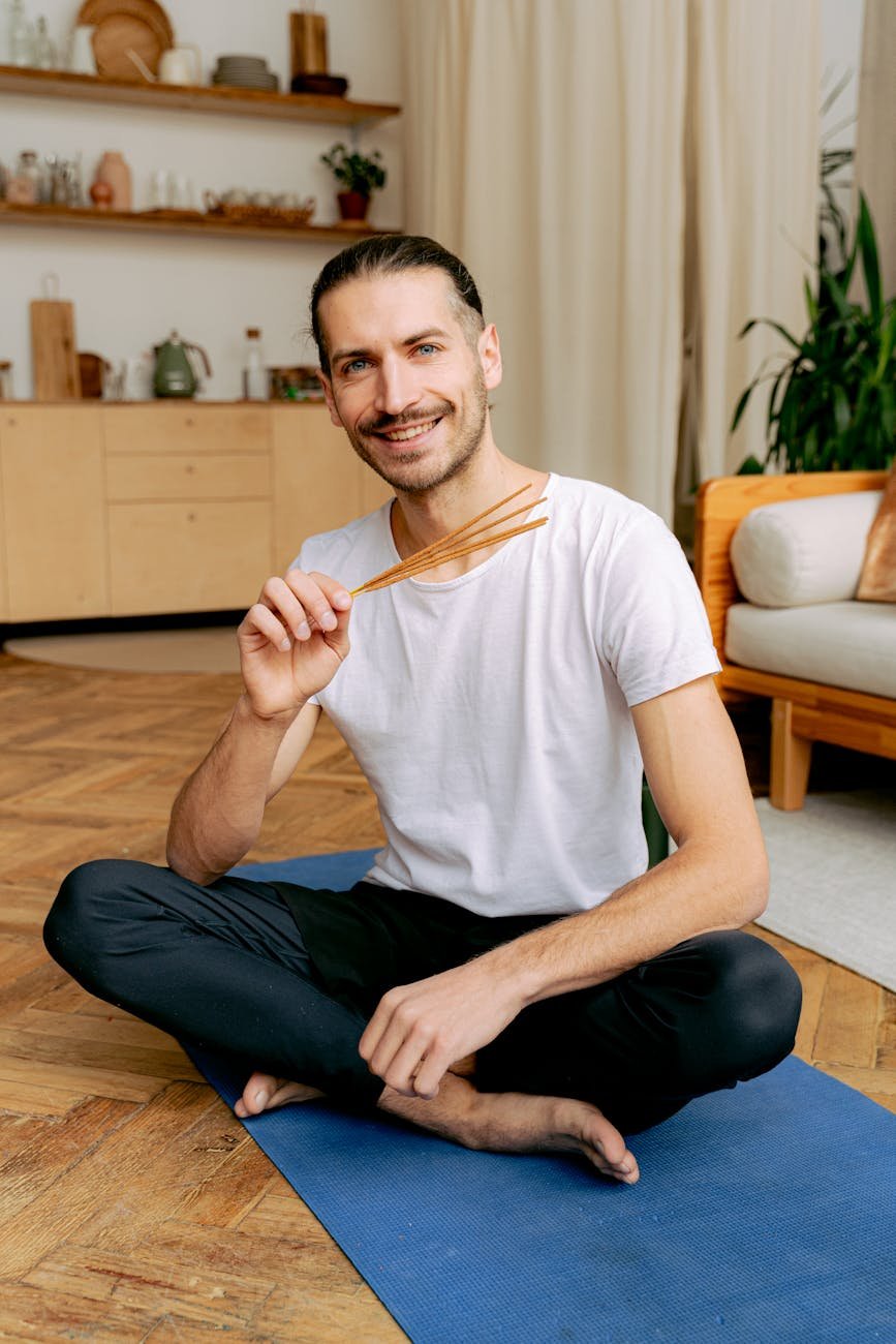 a man in white shirt sitting on his yoga mat while holding an incense sticks